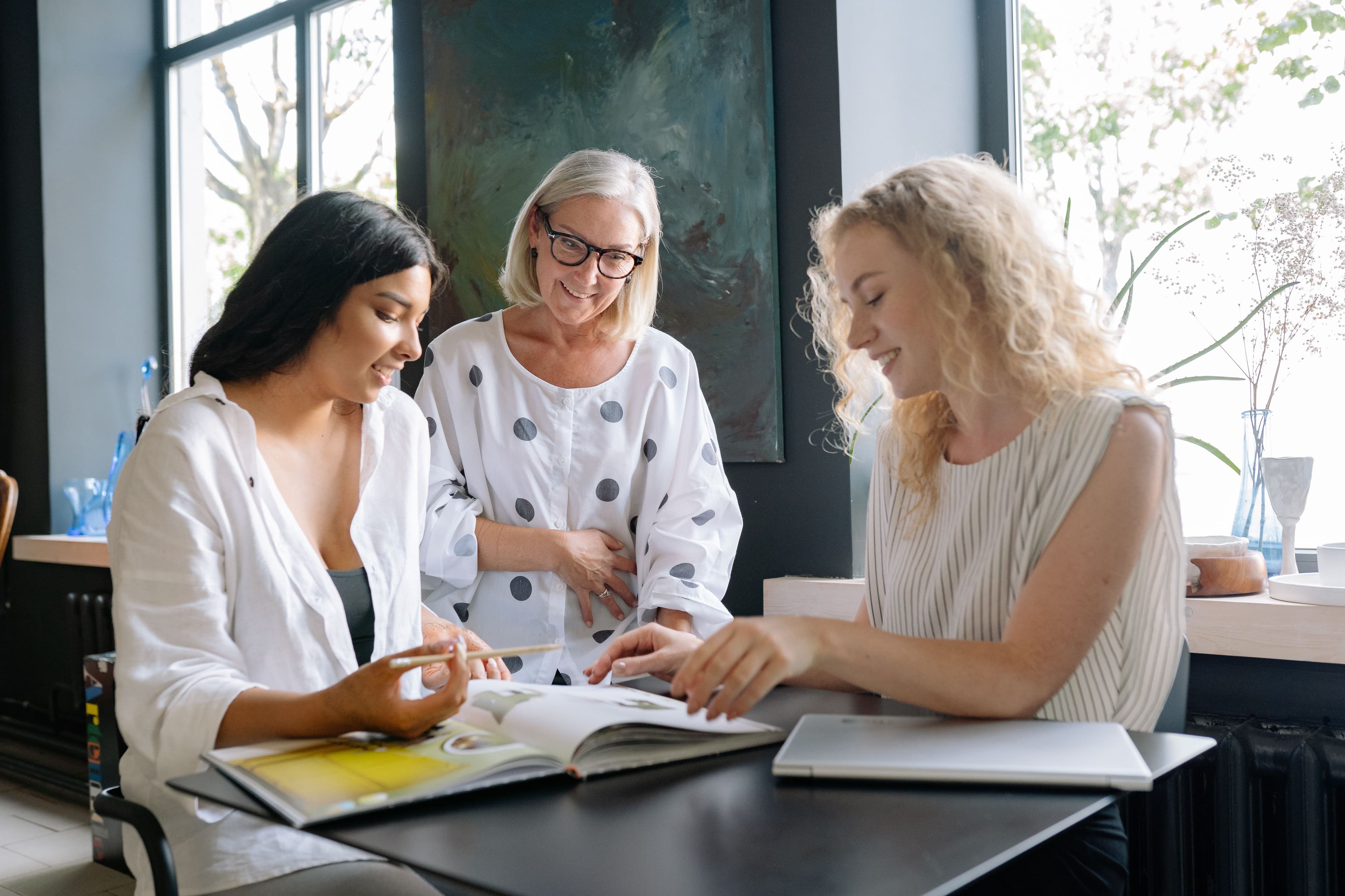 Three women looking through a book together at a table, warm natural light