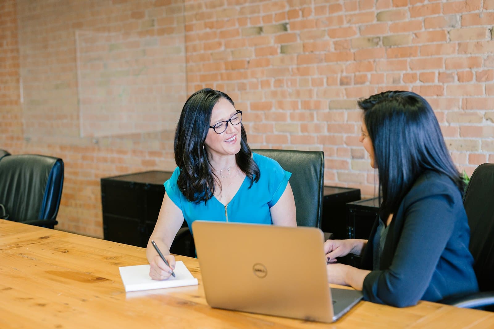 Two women talking and taking notes together at a table against an exposed brick wall