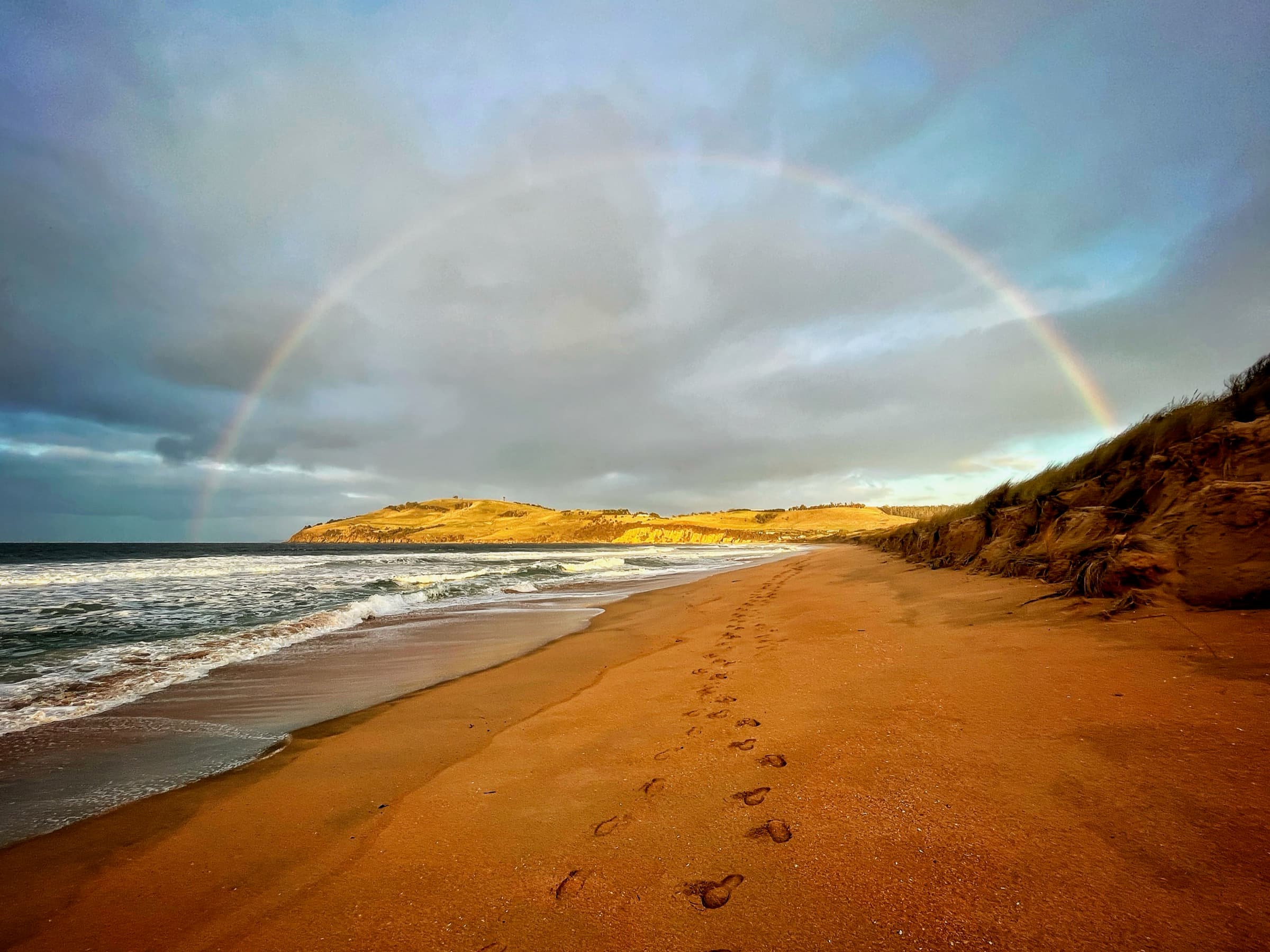 A rainbow arching over a beach, with a trail of footprints leading through the sand